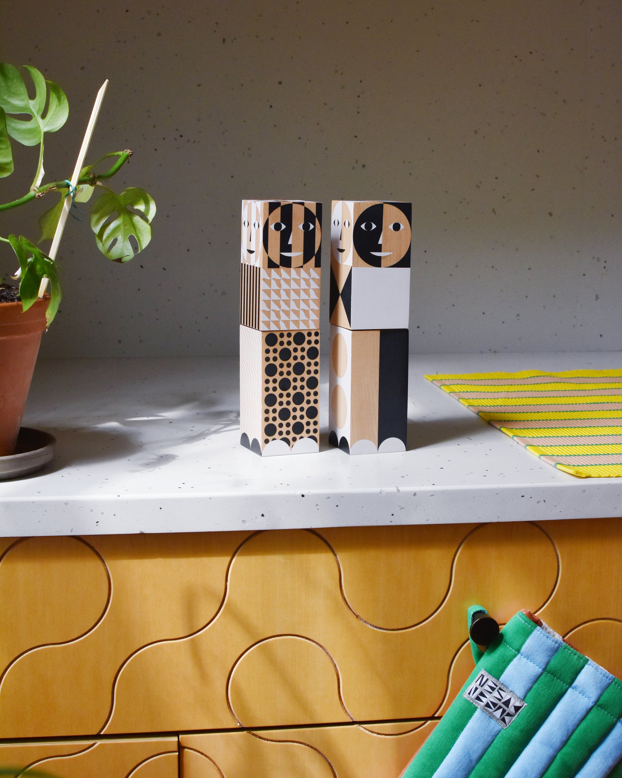 A set of two black and white pepper grinders sit on a kitchen counter top with a blue and green oven mitt and yellow stripe placemat nearby.