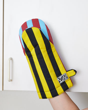 Dusen Dusen Oven Mitt in Mustard (yellow, black, marroon, and blue). Padded oven mitt in varying stripe weights and contrasting stripe colorways. Features a "frog-style" mitt shape, top stitched brand label, and a side loop. 100% cotton quilted canvas base, with polyester fill.