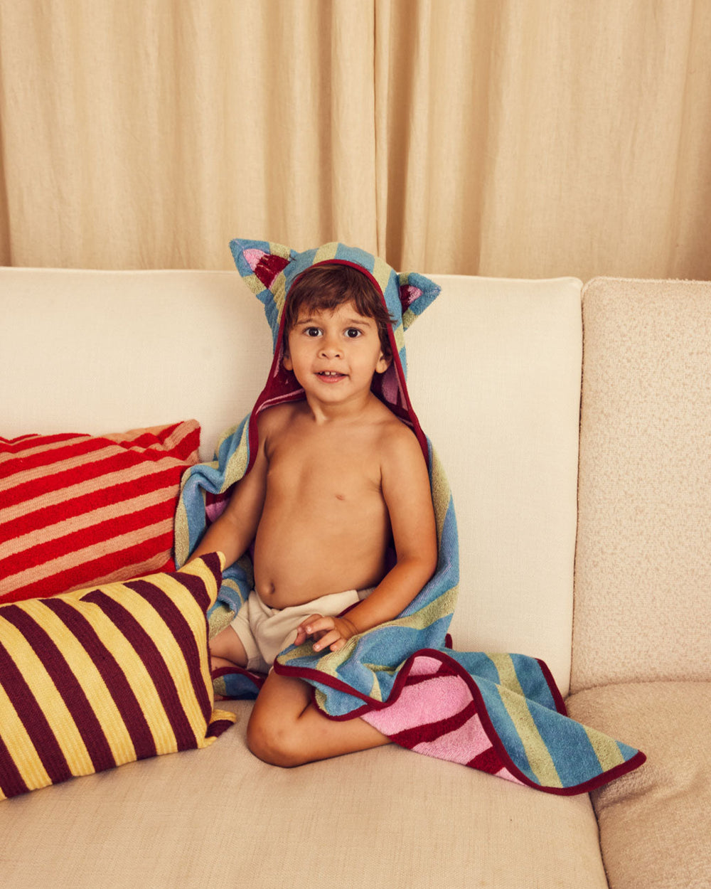 Child sitting on a couch with a colorful towel draped over them, surrounded by striped pillows.