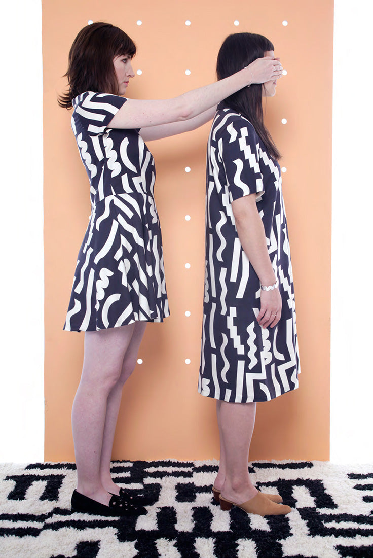 model covering the eyes of the model in front of her both in black dresses with white squiggle pattern
