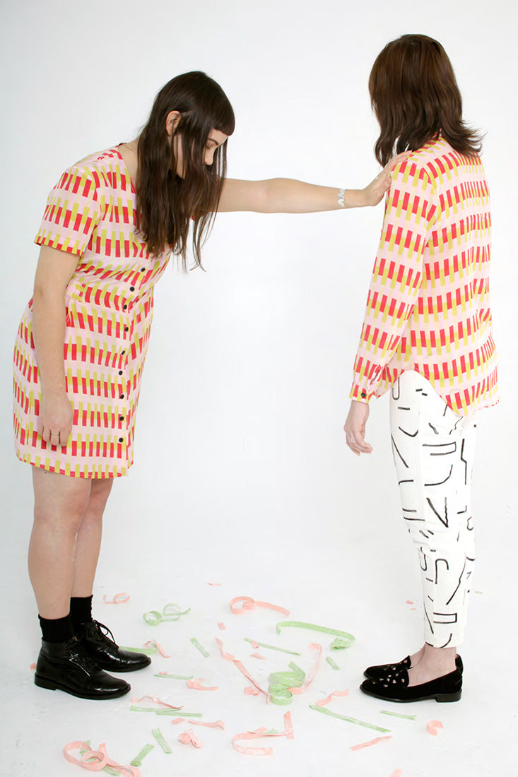 model in button up pink, red and yellow dress putting hand on should of person in pink, red and yellow shirt with white pattern pants