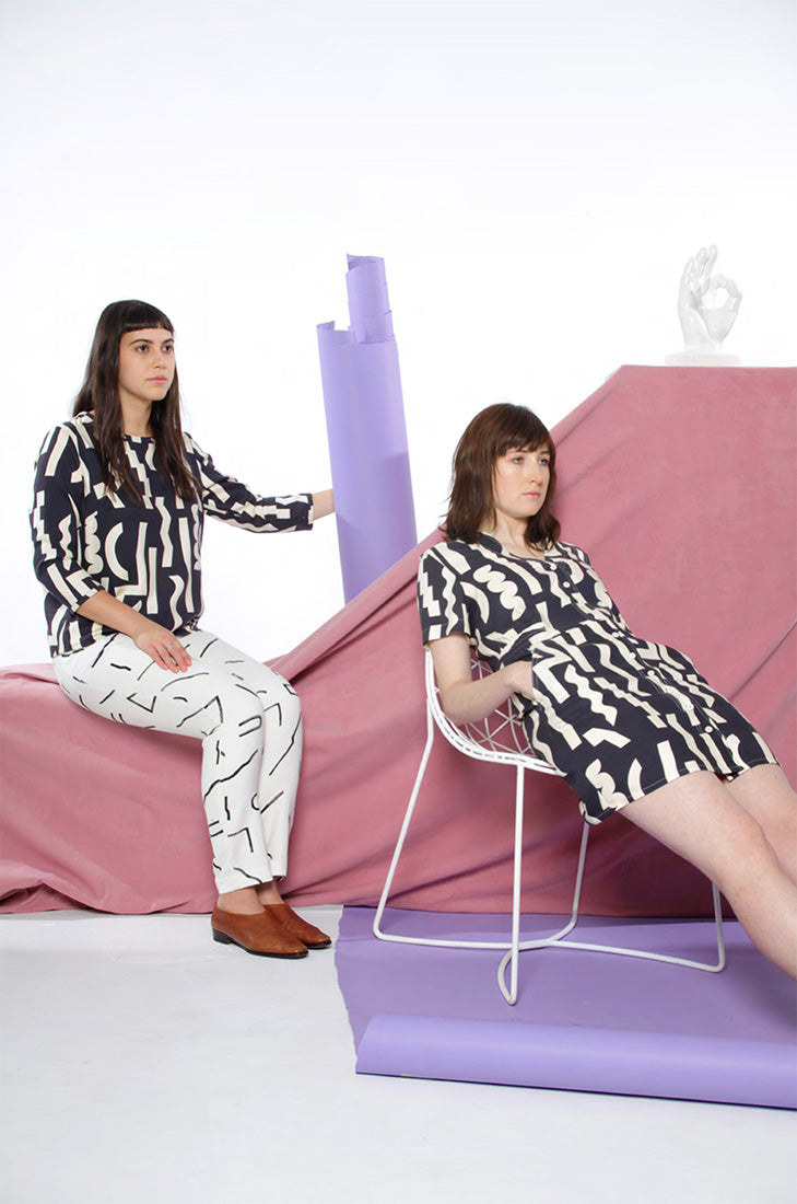 two models sitting, one in hair in black romper with white squiggles and other on a block with black pattern top with white line pattern pants