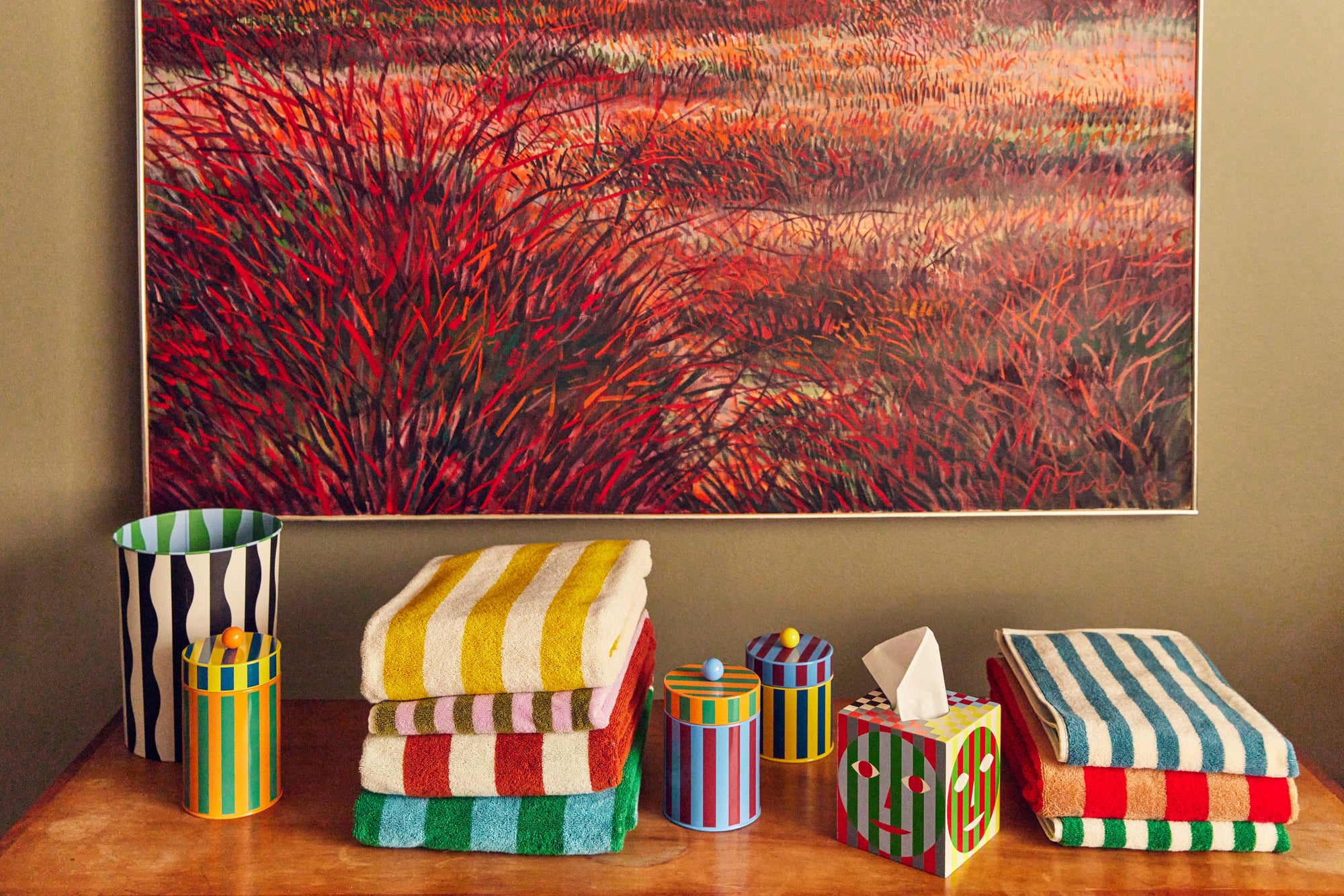 Tableau of Dusen Dusen bath towels, Dusen Dusen canisters, and Dusen Dusen tissue box sit on top of a bench in front of a red field painting