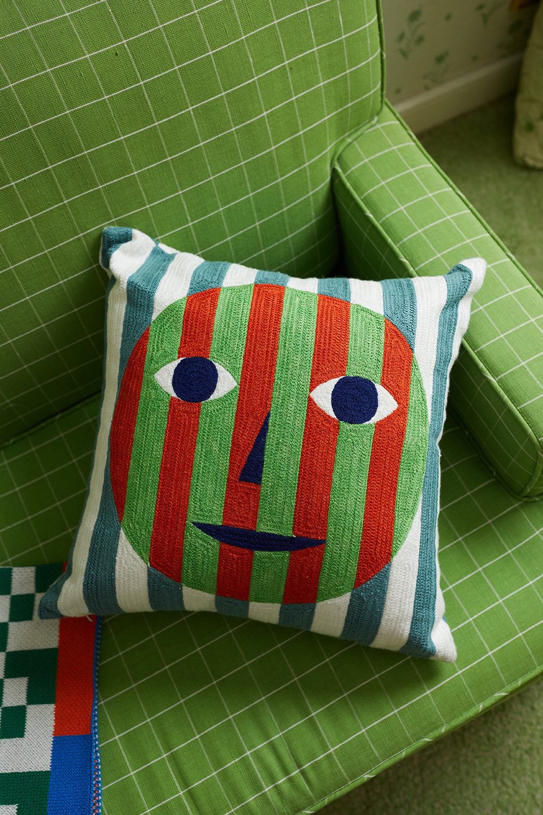 Dusen Dusen embroidered cotton Everybody Face pillow lays on a green checkered couch in a living room