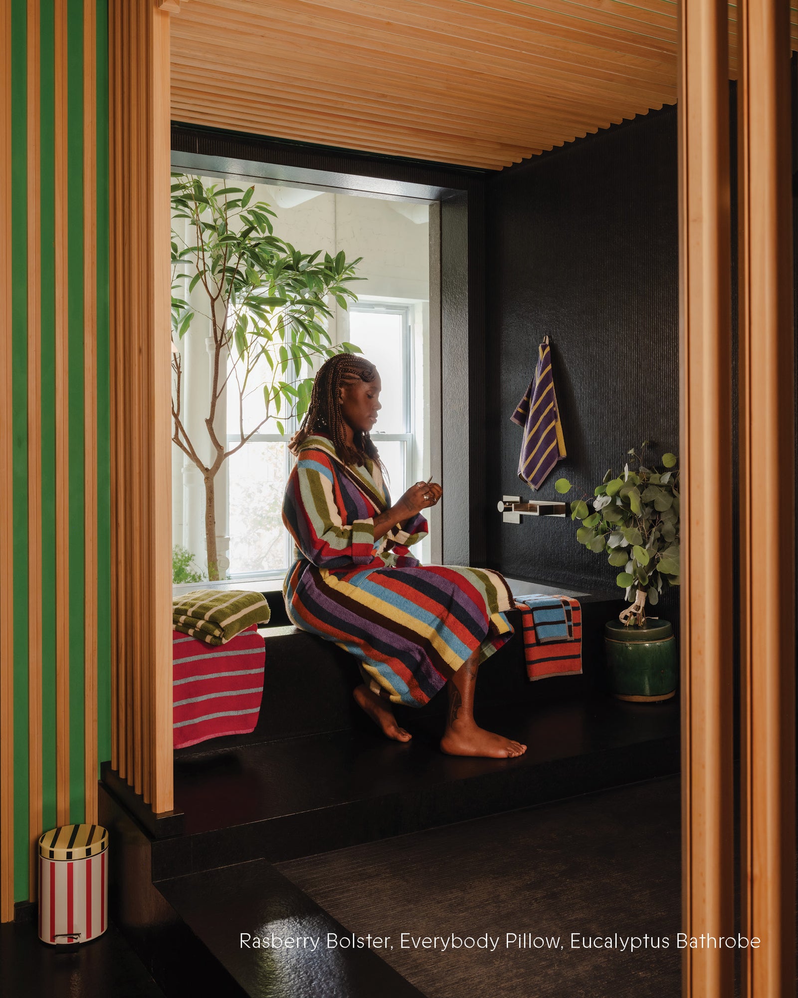 Woman in a colorful striped robe sitting on a bench in a room with striped walls and plants.