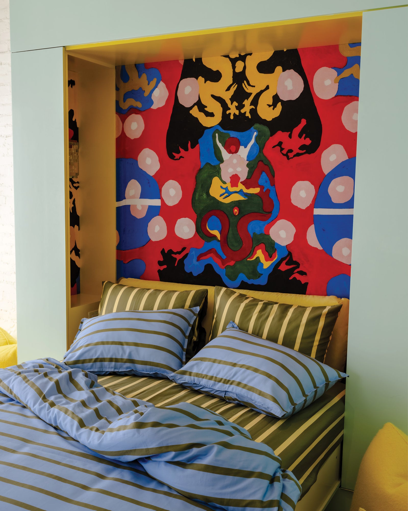Bedroom with colorful wall art and Cornflower striped bedding, next to a room with hanging striped bathrobes and marigold hamper on the floor with slippers