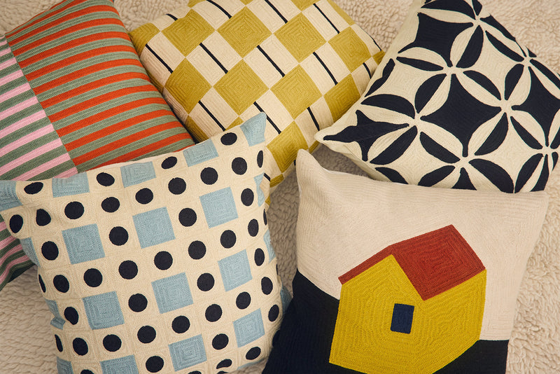A group of five decorative throw pillows with various colorful patterns, including stripes, checkers, polka dots, and a house design, arranged together