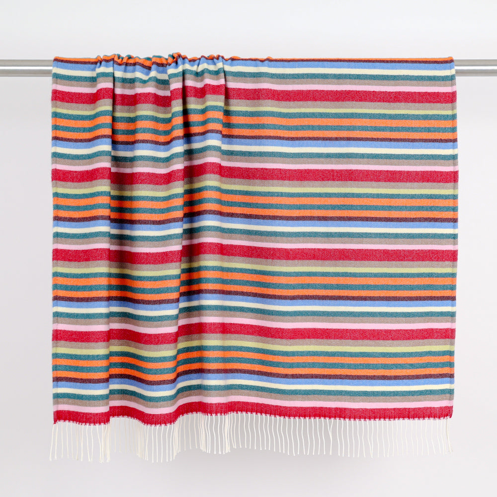 A multicolor striped wool blanket with white fringe, hanging from a metal rod against a plain white wall.