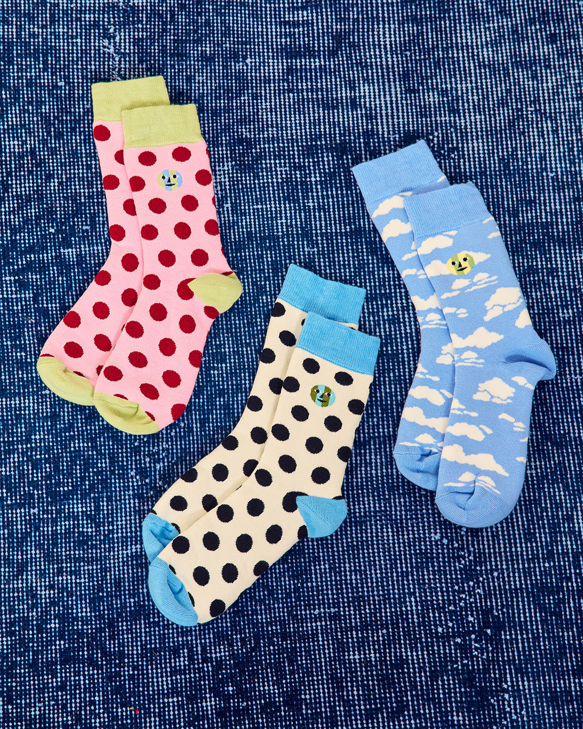 Three pairs of patterned crew socks arranged on a textured blue background. One pair is pink with red polka dots and light green cuffs/soles, one pair is cream with black polka dots and light blue cuffs/soles, one pair is light blue with white cloud patterns and a small yellow smiley face. 