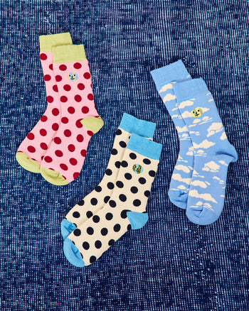 ["Three pairs of patterned crew socks arranged on a textured blue background. One pair is pink with red polka dots and light green cuffs/soles, one pair is cream with black polka dots and light blue cuffs/soles, one pair is light blue with white cloud patterns and a small yellow smiley face. ", " You'll have lots to trade on the New York Sock Exchange"]
