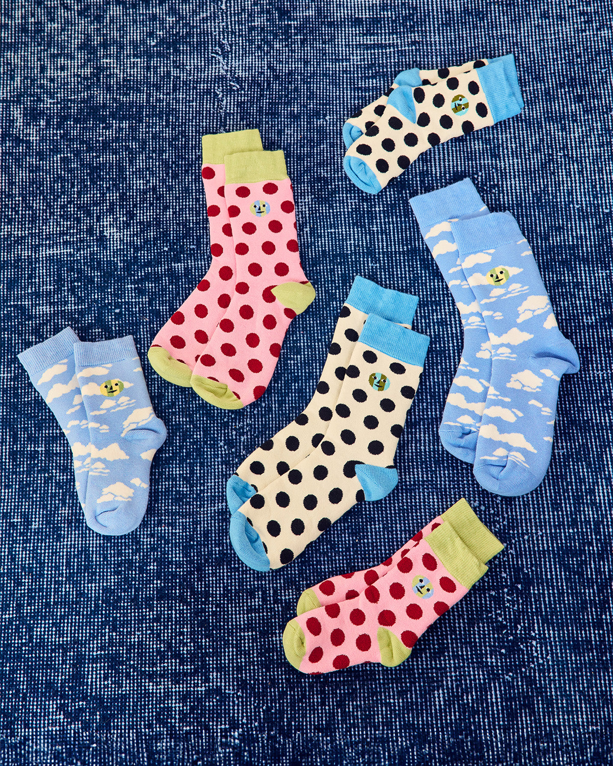 Six pairs of children's socks in various colorful patterns (polka dots, red strawberries, clouds, and black spots on cream) are arranged on a textured blue background.