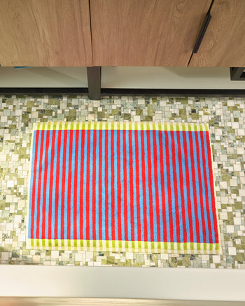 A rectangular rug with vertical red, blue, and lime green stripes, placed on a mosaic tile floor in front of a wooden cabinet. 