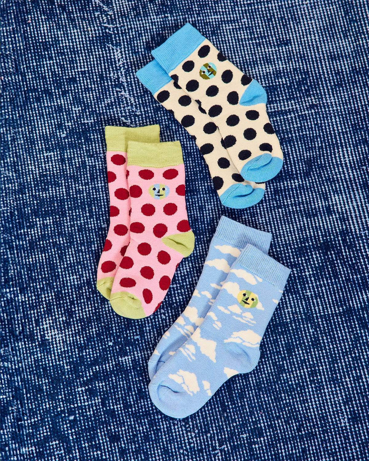 Three pairs of patterned baby socks arranged on a blue denim-like fabric background. One pair is cream with black polka dots and bright blue toes/heels, one pair is pink with red polka dots and light green toes/heels. one pair is light blue with white cloud patterns and yellow-green accents. 