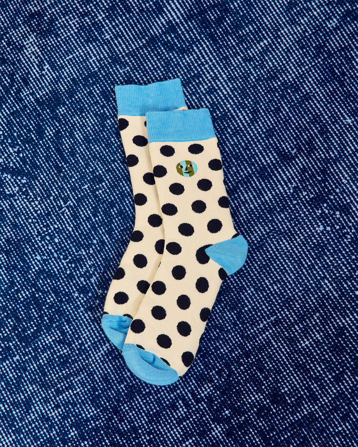 A pair of cream-colored socks with black polka dots, light blue cuffs, heels, and toes, featuring a small embroidered logo on the upper leg, laid flat on a blue denim background. 