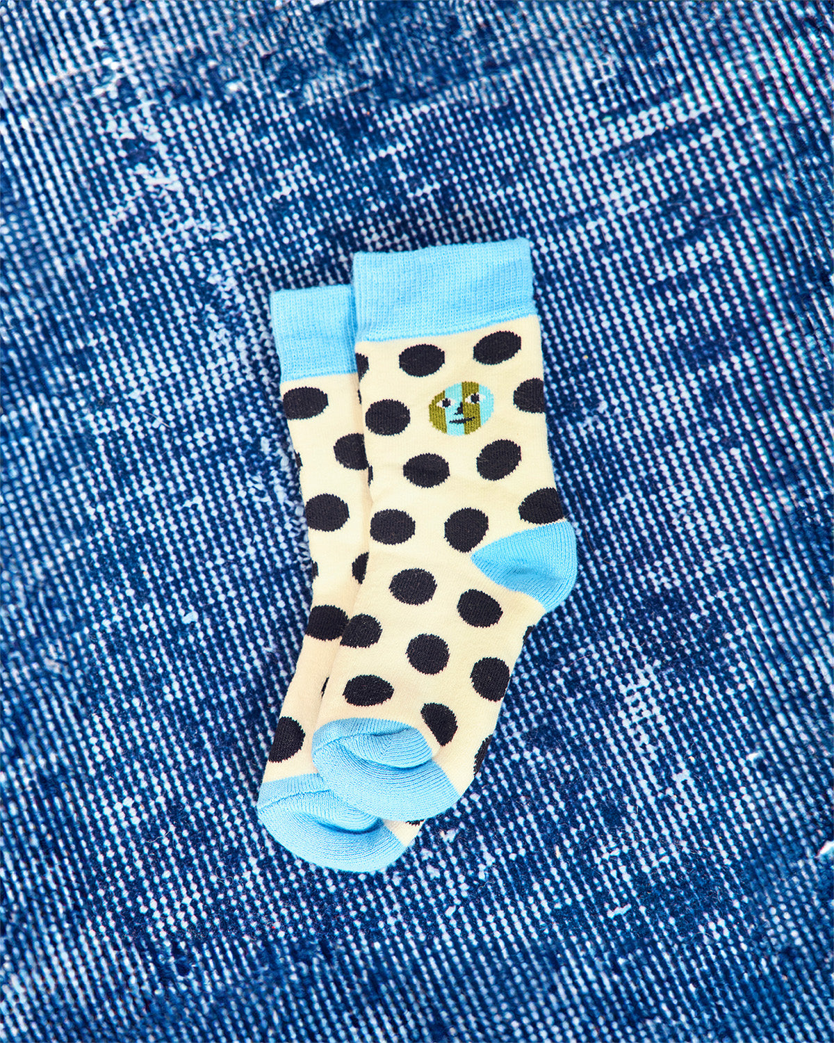 A pair of cream-colored socks with black polka dots, light blue cuffs and heels, and a small circular graphic on the side, laid flat on a blue denim background.
