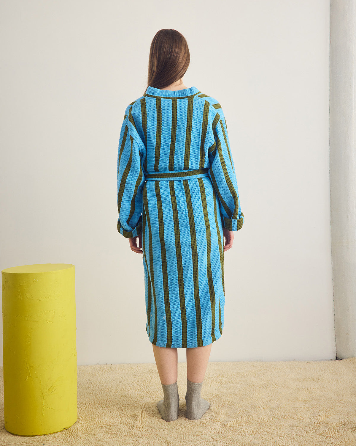 Person wearing a blue and green striped robe standing in a minimalistic room with a yellow cylindrical object.
