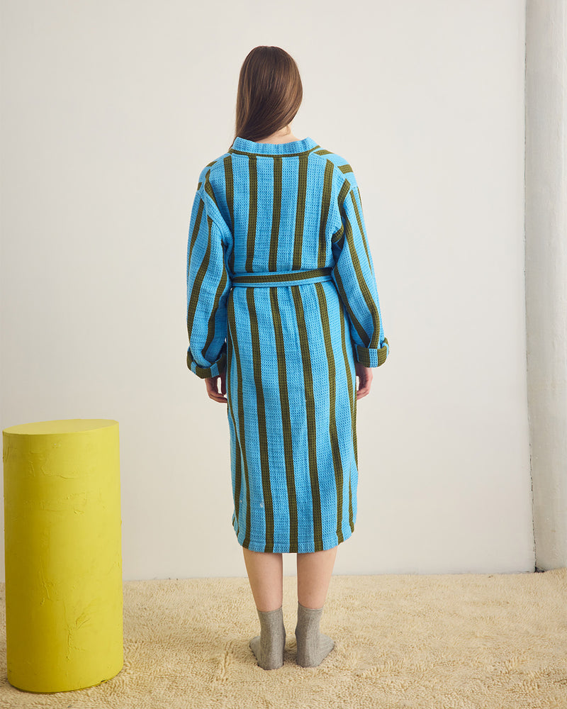 Person wearing a blue and green striped robe standing in a minimalistic room with a yellow cylindrical object.