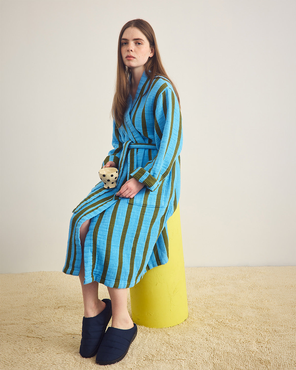 Woman wearing a blue and green striped dress with a yellow vase in a minimalistic setting