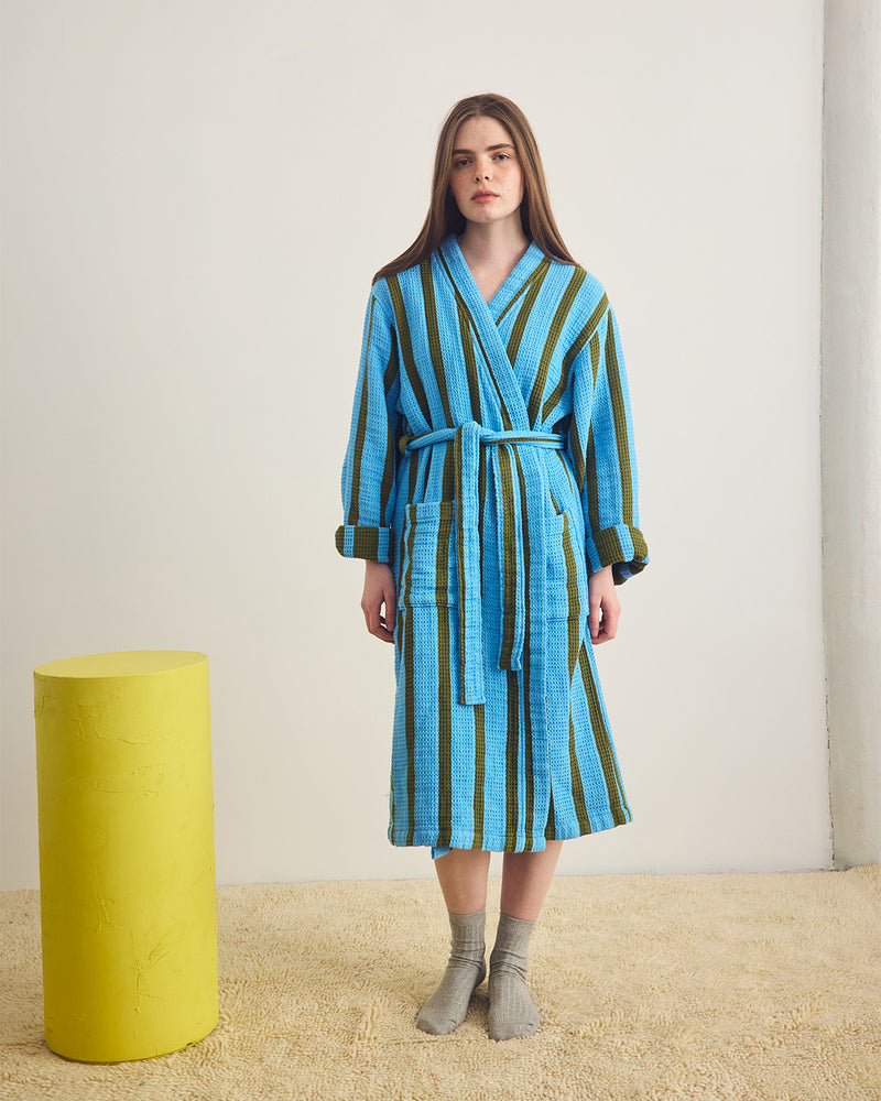 Woman wearing a blue and green striped robe standing next to a yellow cylindrical object on a beige carpet.