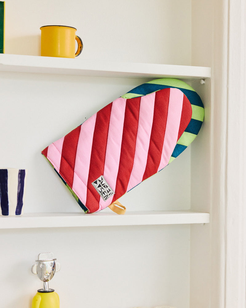 Colorful striped oven mitt on a white shelf with a yellow mug and other items. 
