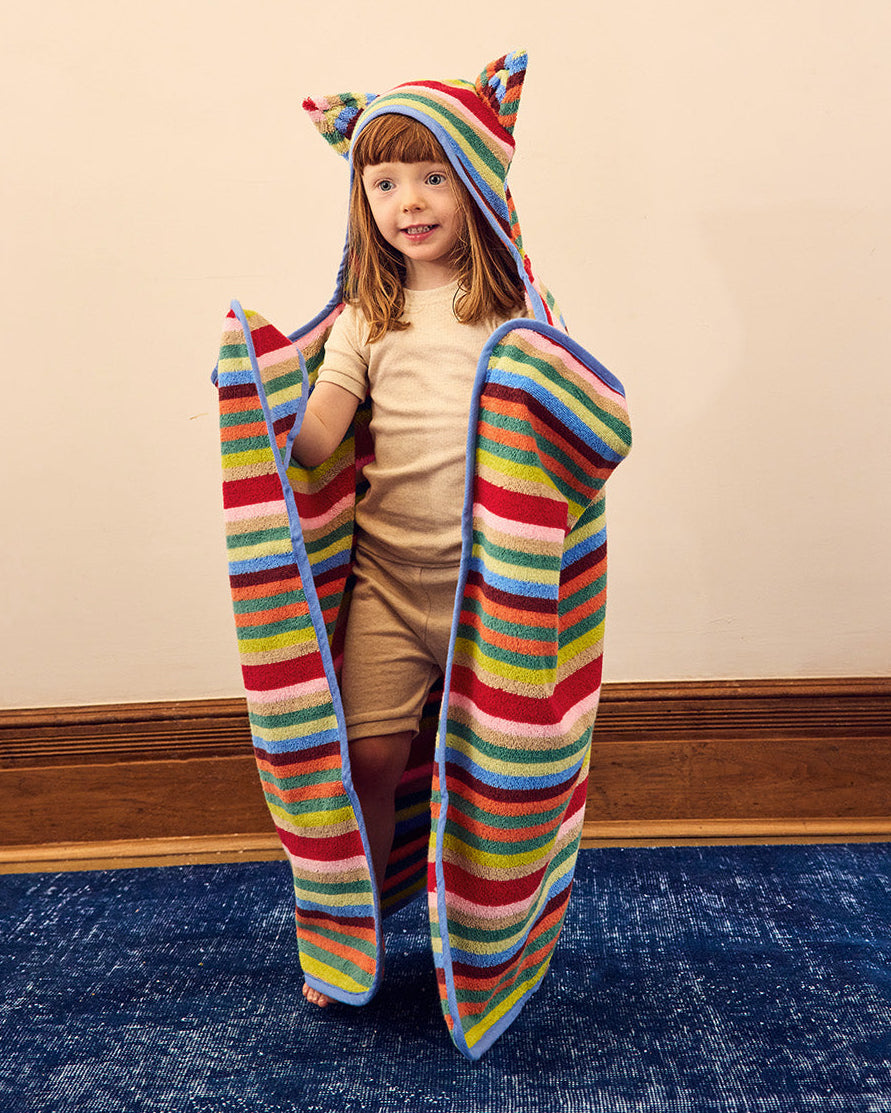 A young child wearing a colorful striped hooded blanket with ears stands on a blue carpeted floor against a beige wall. 