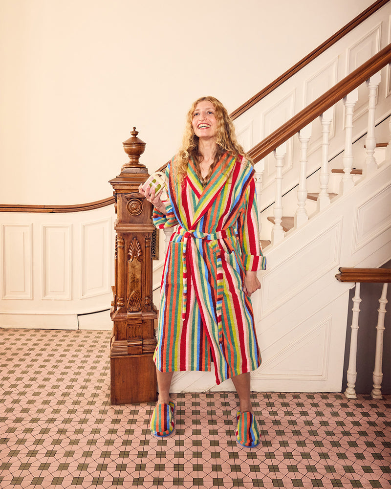 A woman stands in a grand staircase hall wearing a colorful vertically striped bathrobe and matching striped slippers. The robe features bold stripes of red, blue, green, yellow, and white, with a matching tie belt.