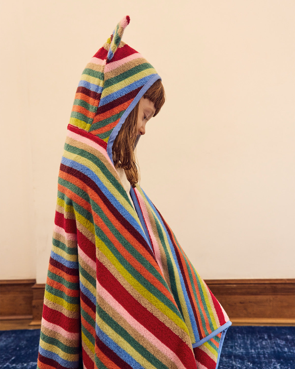 A child wrapped in a colorful striped hooded towel , standing indoors against a plain beige wall with wood trim and a blue rug visible.