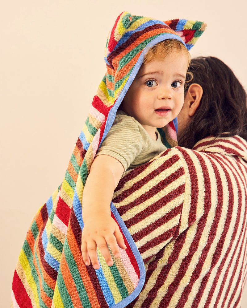 A baby wrapped in a colorful striped hooded towel with cat ears, held by an adult wearing a matching striped bath towel, against a plain beige background.