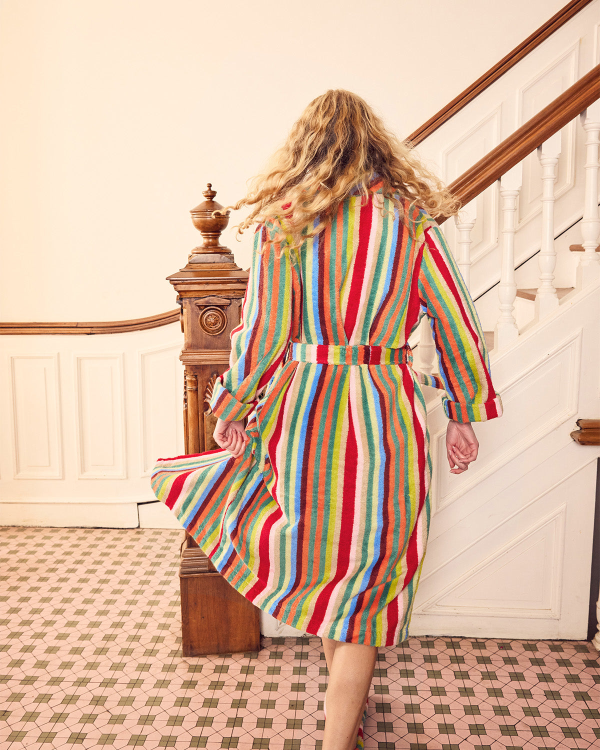 A woman viewed from behind wearing a long-sleeved, belted striped dress with vertical stripes in red, green, blue, yellow, and white, walking down a staircase in an indoor setting.