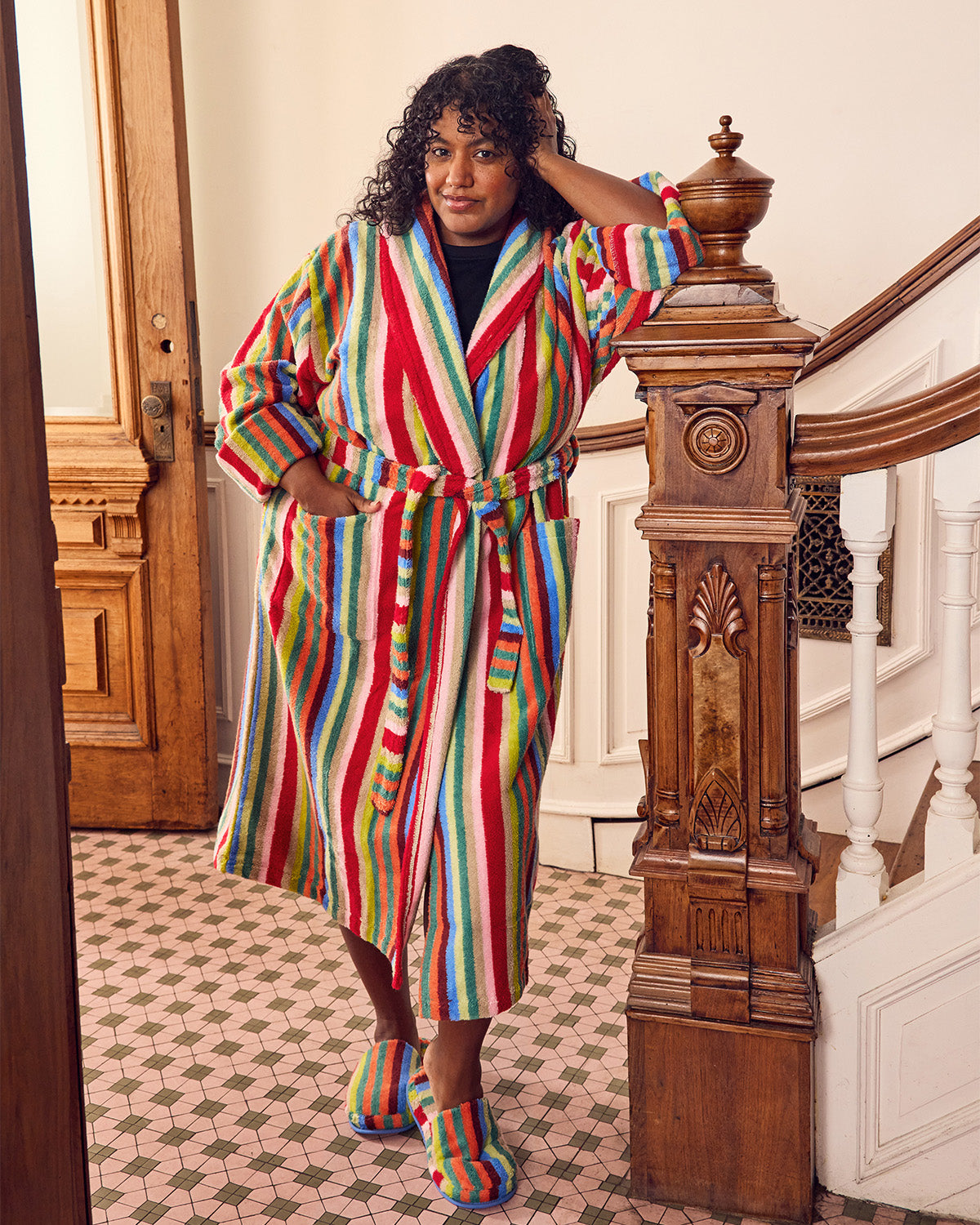 A woman stands in a staircase hallway wearing a long, colorful vertically striped bathrobe with matching striped slippers. The robe features bold stripes in red, green, blue, yellow, and white, and she is posed next to a carved wooden banister.