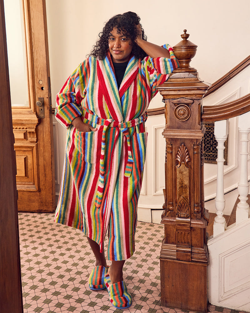A woman stands in a staircase hallway wearing a long, colorful vertically striped bathrobe with matching striped slippers. The robe features bold stripes in red, green, blue, yellow, and white, and she is posed next to a carved wooden banister.