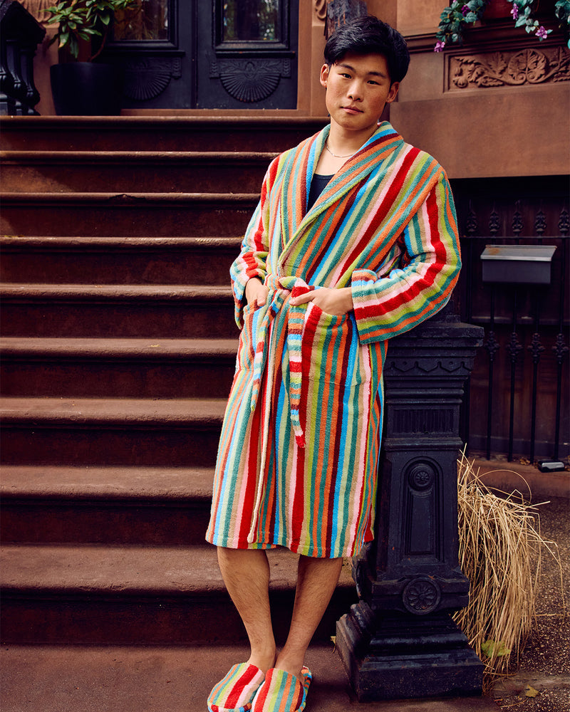 A man wearing a colorful vertically striped bathrobe with red, blue, green, yellow, and white stripes, standing on outdoor stone steps in front of a brown building entrance.