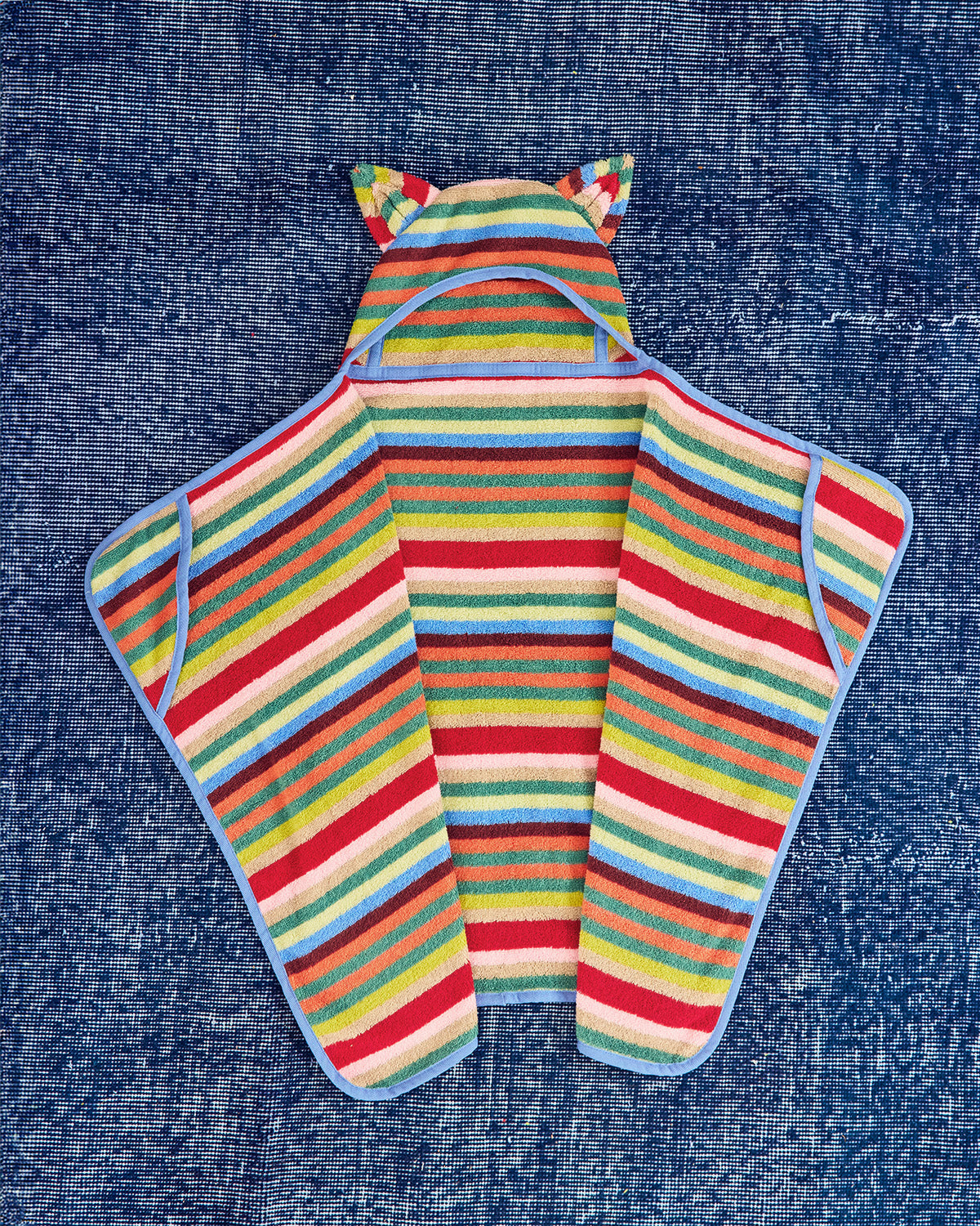 A colorful striped hooded towel laid flat on a blue textured fabric background. The hood has cat-ear details, and the body features horizontal stripes in red, yellow, green, blue, pink, and white, with a light blue border.