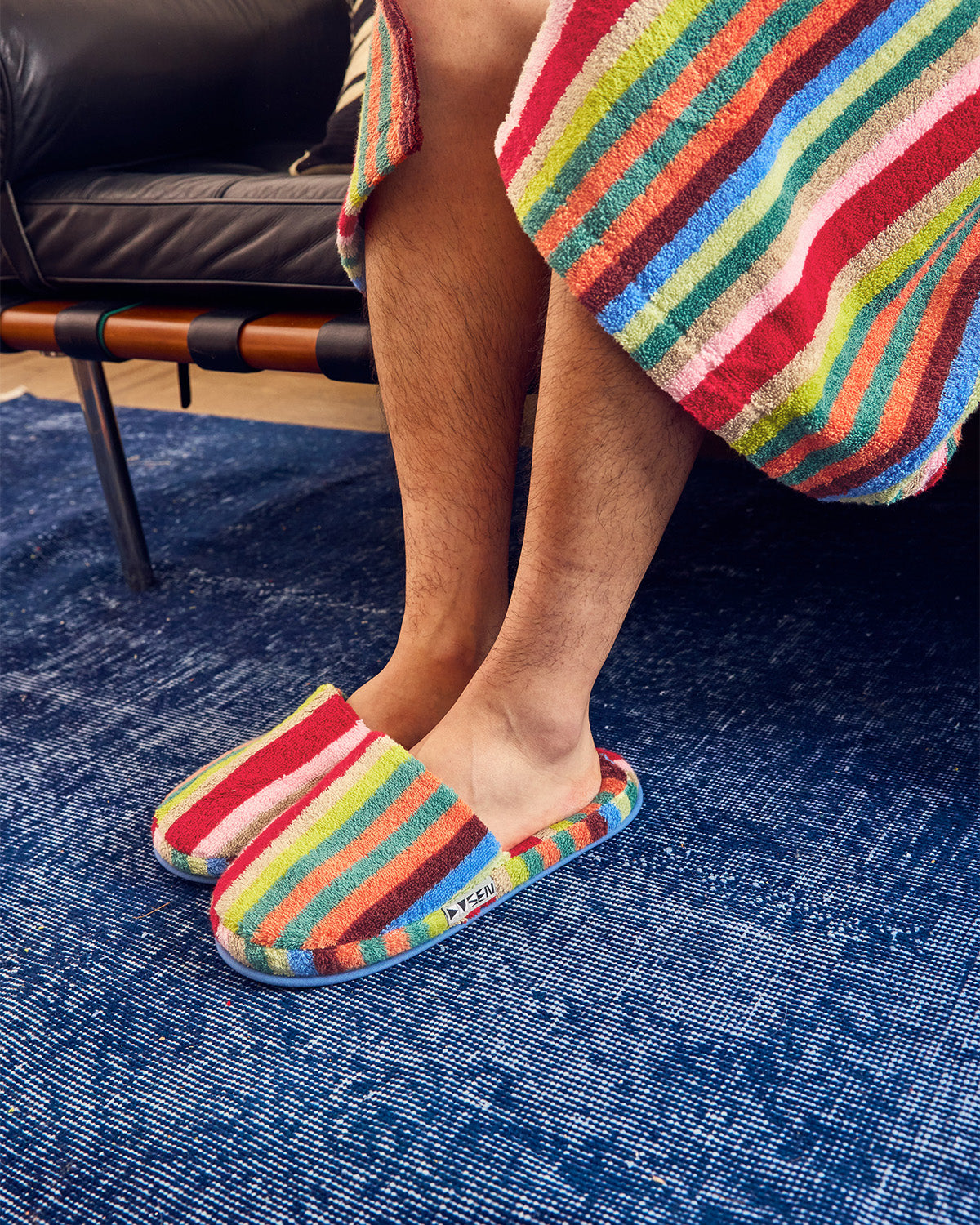 A person sitting indoors wearing colorful striped slip-on slippers and a matching striped towel draped over their legs, with a blue textured rug and dark leather furniture in the background. 