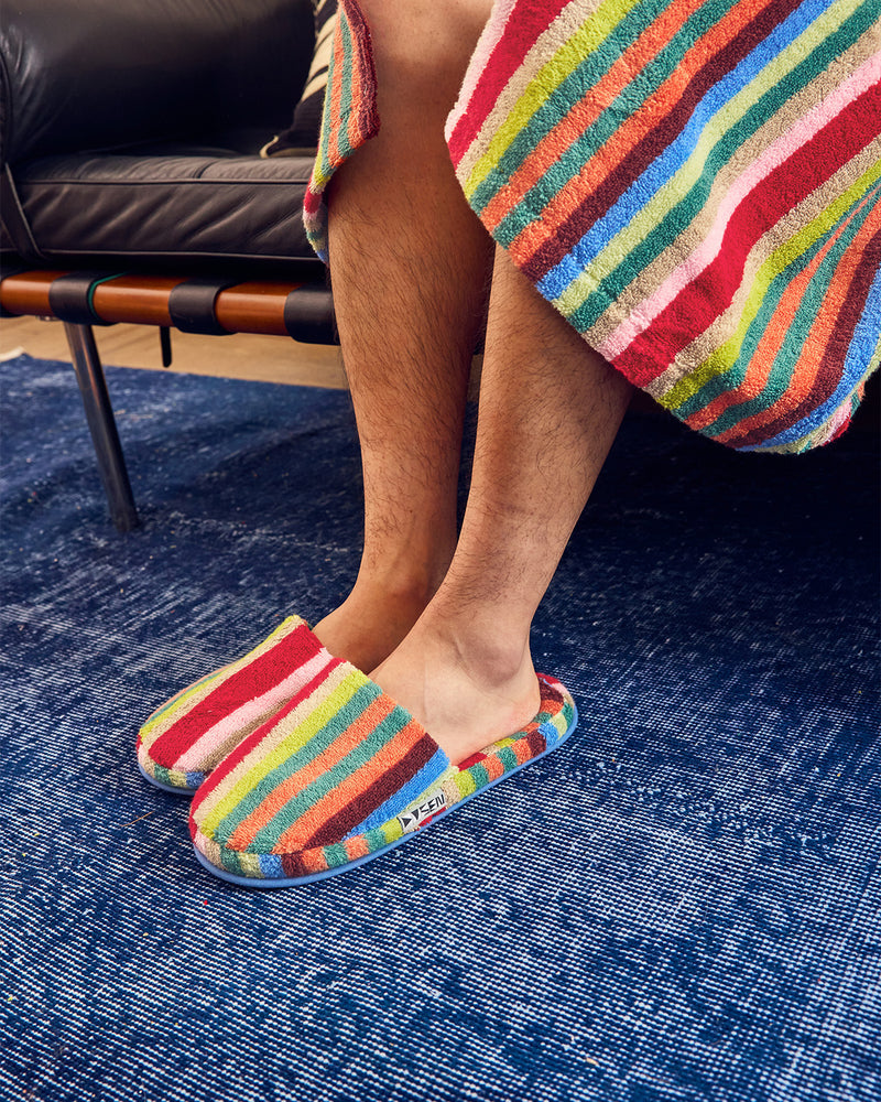 A person sitting indoors wearing colorful striped slip-on slippers and a matching striped towel draped over their legs, with a blue textured rug and dark leather furniture in the background. 