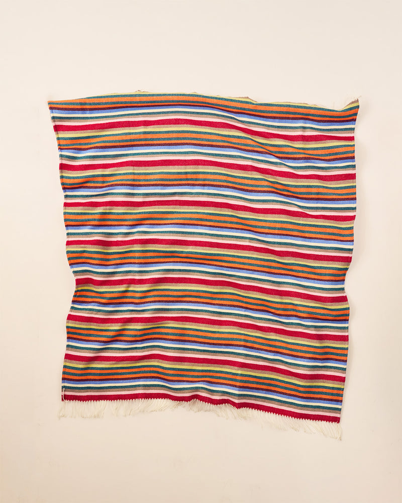 A merino wool throw with horizontal multicolor stripes in red, orange, yellow, green, blue, and white, finished with short fringe on one edge, laid flat against a light beige background. 