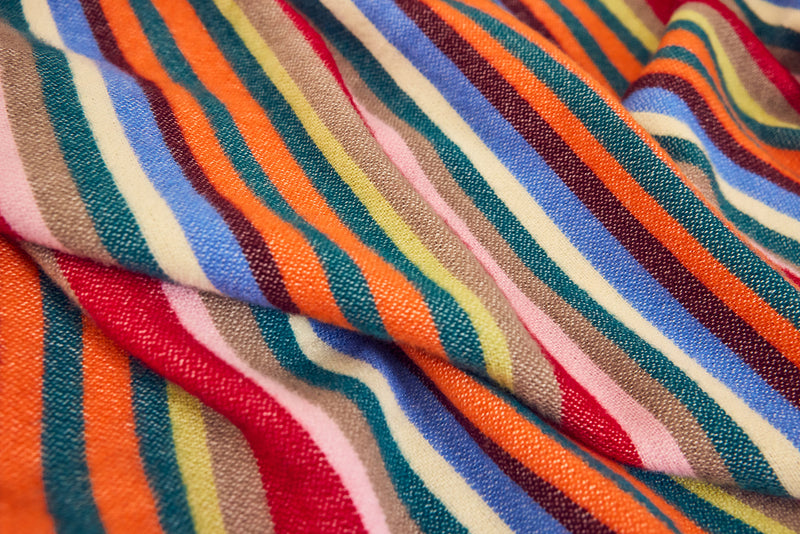 A close-up of a merino wool with woven vertical bands in orange, red, pink, cream, blue, teal, brown, and dark red, shown with soft folds.