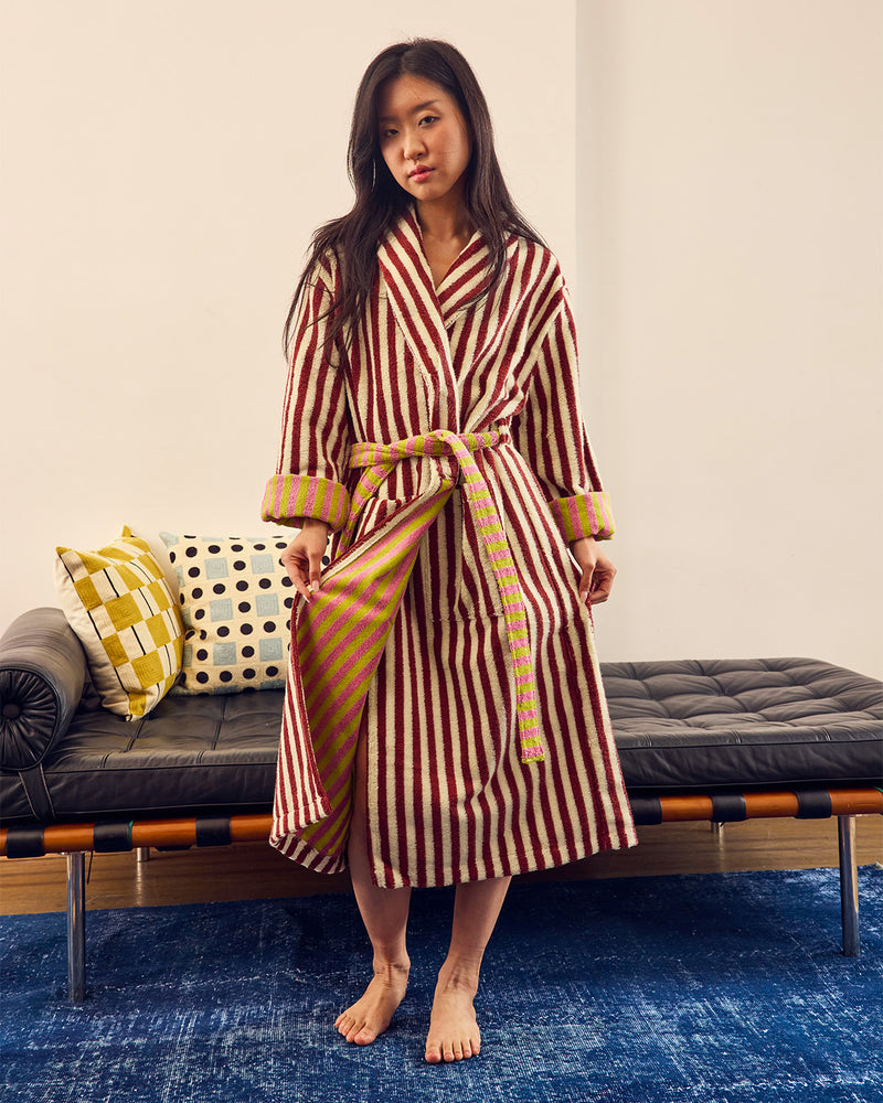 A woman stands barefoot in a bedroom setting wearing a long striped bathrobe with a tie belt. The robe has vertical red and cream stripes with yellow and green patterned cuffs and hem.
