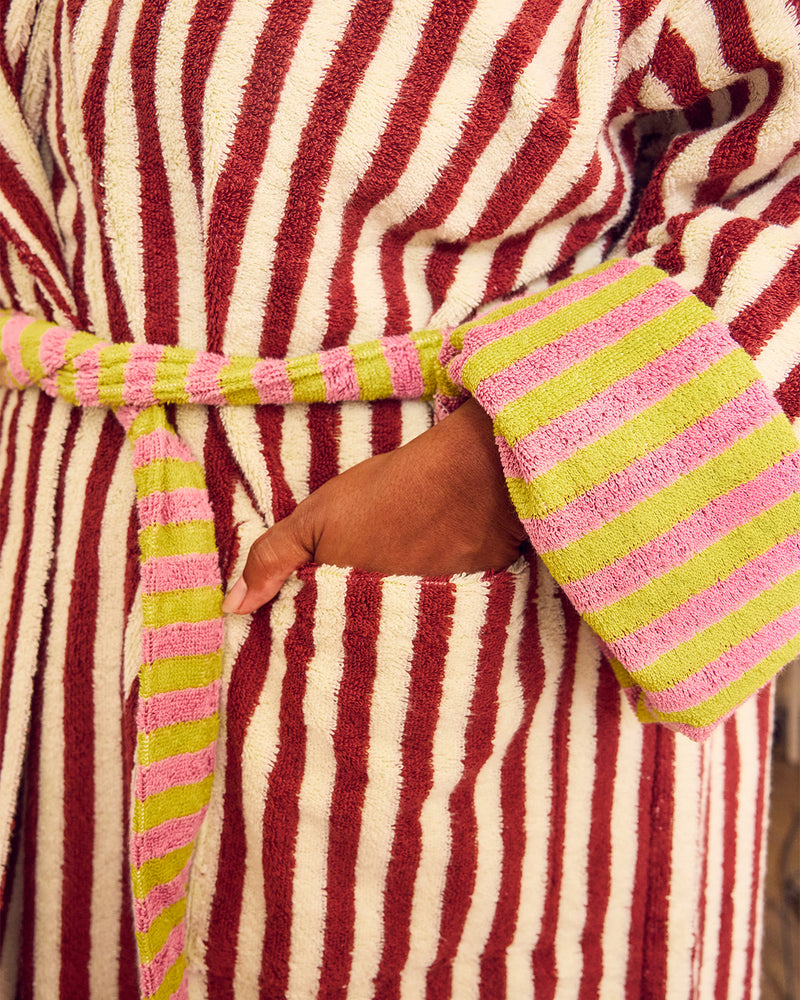 A close-up of a striped bathrobe with a hand in a front pocket, featuring vertical red and cream stripes with pink and lime green striped cuffs and waist tie.