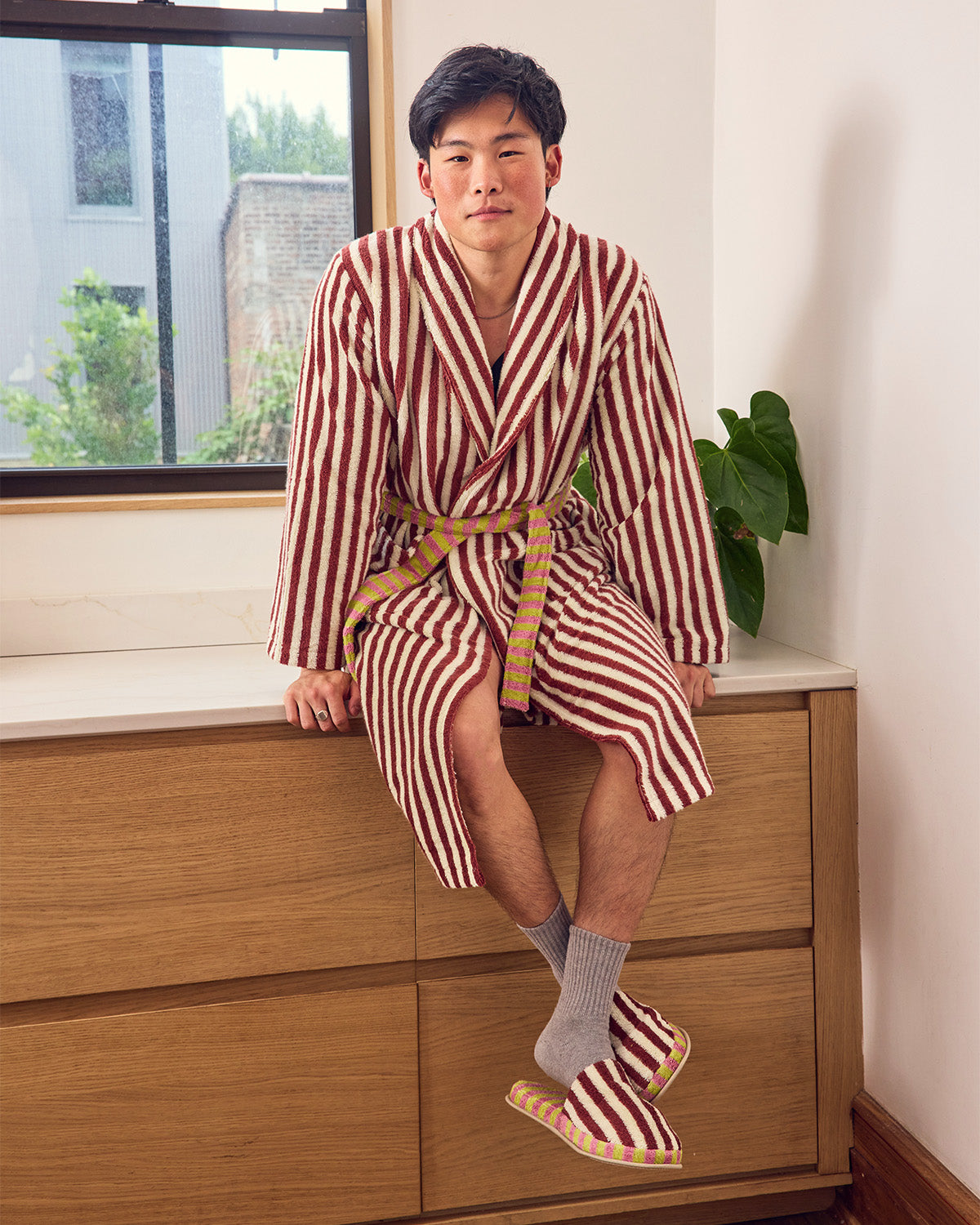 A man wearing a maroon and white vertically striped bathrobe with a tan belt, gray socks, and matching striped slippers, sitting on a wooden dresser in a bright room.