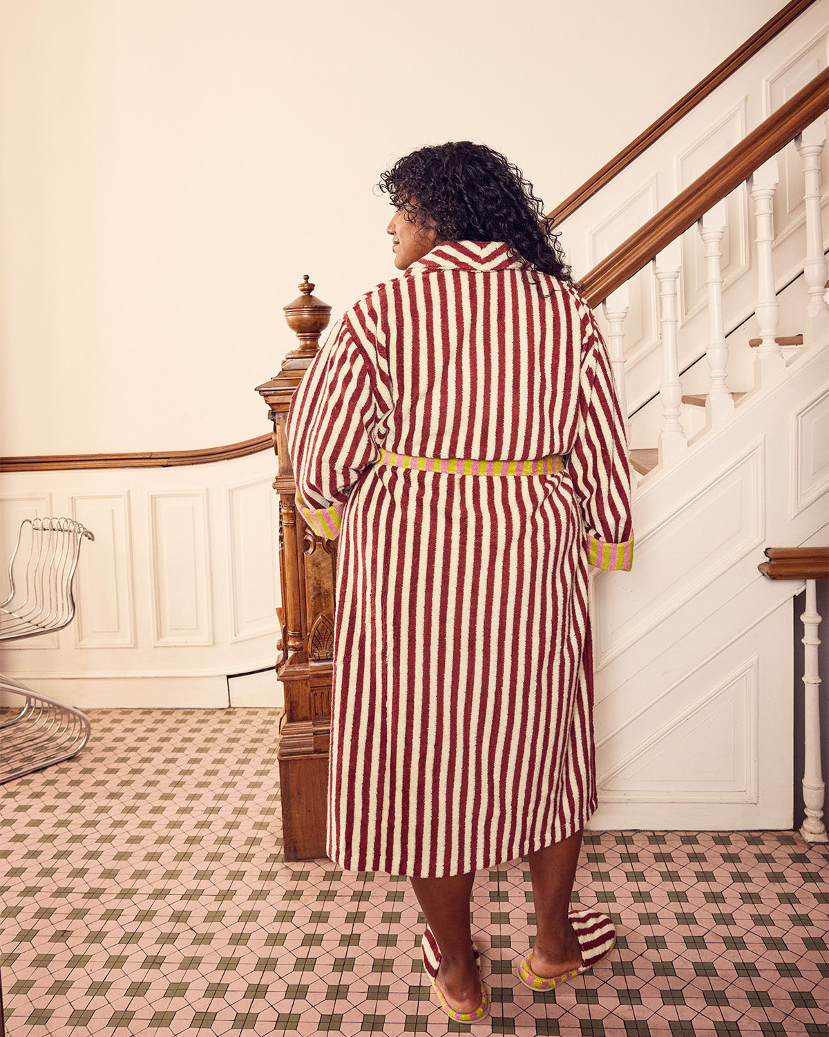 A woman viewed from behind wearing a red and white vertically striped bathrobe with a matching belt, standing in a classic indoor hallway with a staircase.