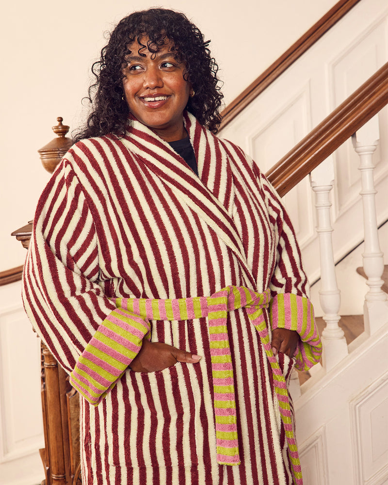 A smiling woman with curly black hair stands on a staircase wearing a plush striped bathrobe with vertical red and cream stripes, a matching tie belt, and striped cuffs in pink and lime green.