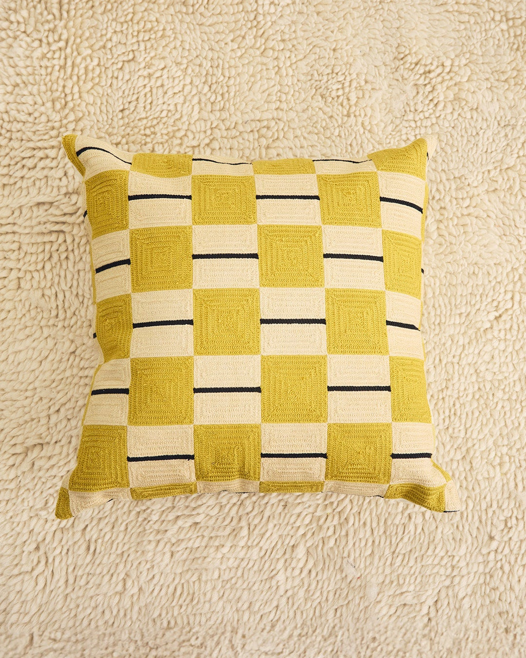 A square throw pillow with a yellow and cream checkerboard pattern featuring black accent stripes, displayed on a textured cream shag rug. 