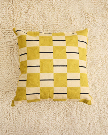 ["A square throw pillow with a yellow and cream checkerboard pattern featuring black accent stripes, displayed on a textured cream shag rug. ", " Simply a pawn in the game of comfort"]