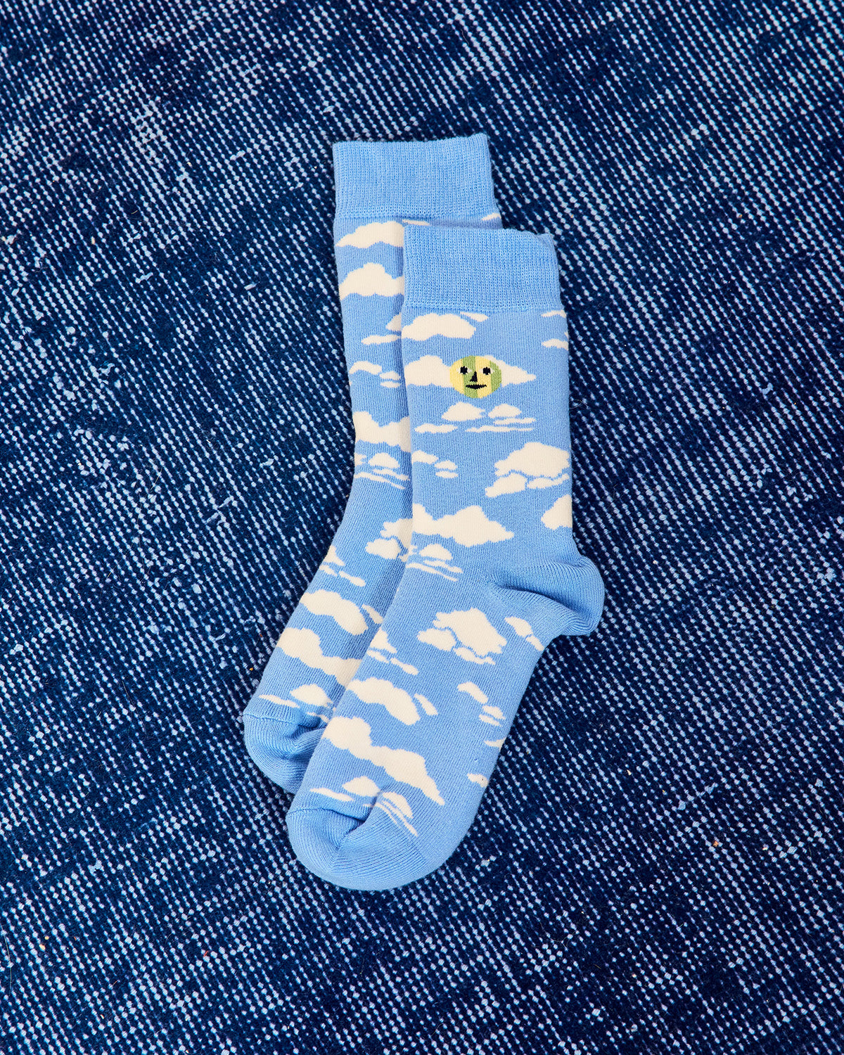 A pair of light blue crew socks with a white cloud pattern and a small yellow smiley face detail, laid flat on a dark blue denim background. 