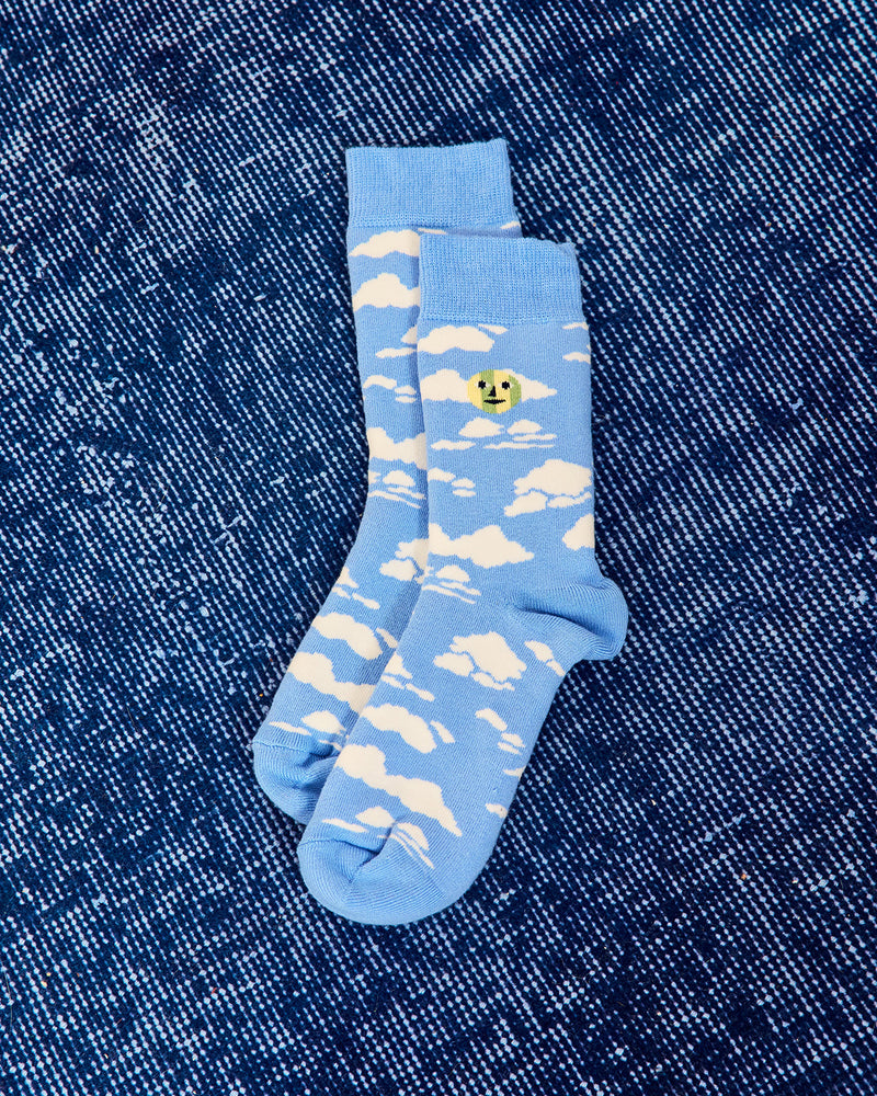 A pair of light blue crew socks with a white cloud pattern and a small yellow smiley face detail, laid flat on a dark blue denim background. 