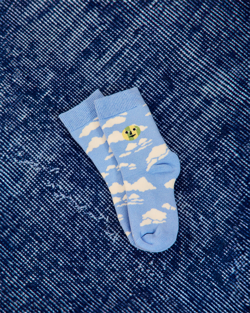 A pair of light blue crew socks with a white cloud pattern and a small yellow smiley face graphic, laid flat on a dark blue denim background.