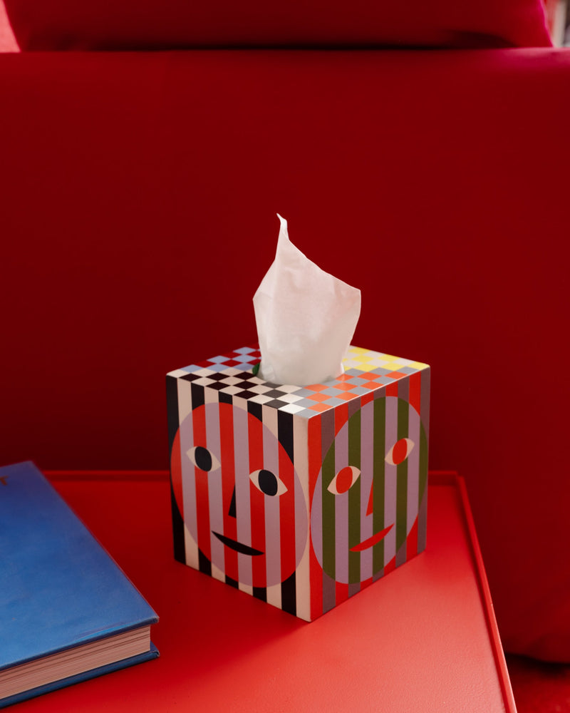 Tissue box holder made in collaboration with Areaware on red table with red couch behind. Printed tissue box holder with signature Face print on each side. Sits over a standard cube tissue box. 100% plywood.