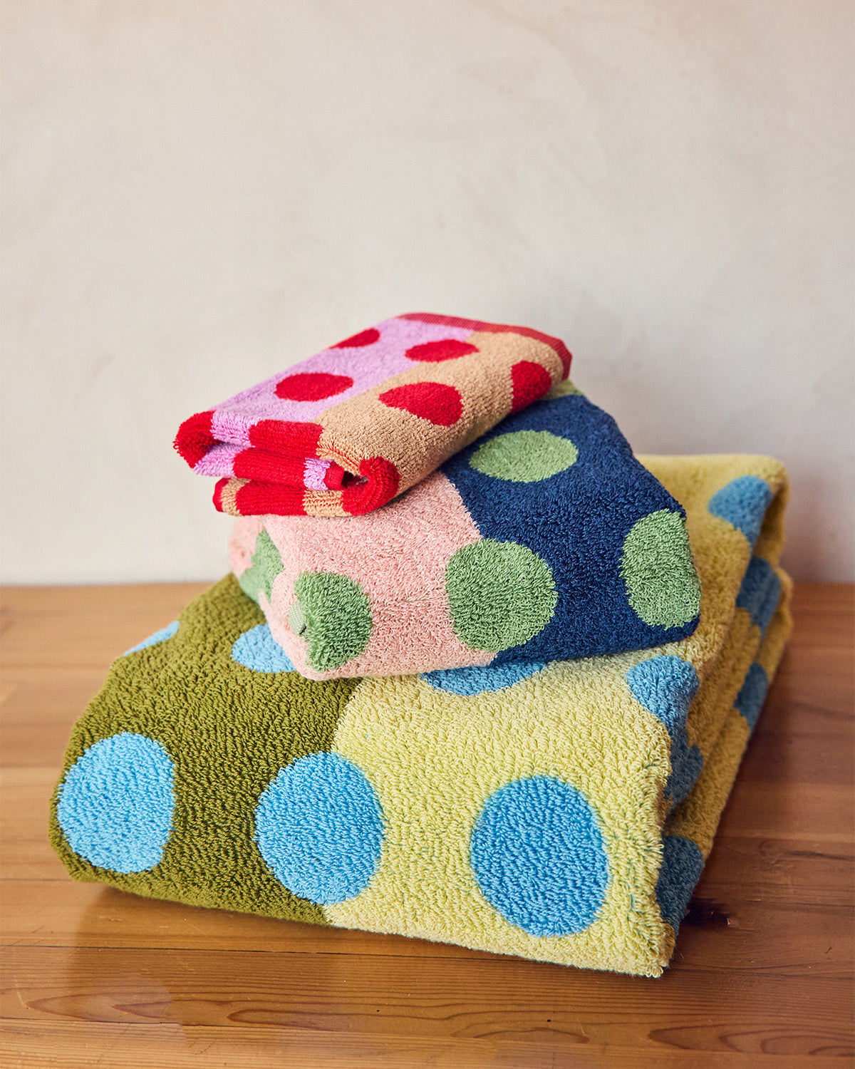 Stack of colorful polka dot towels on a wooden surface with a neutral background <br> <br> Set of 3 includes 1x Blue Dot Bath Towel, 1x Green Dot Hand towel and 1x Red Dot washcloth