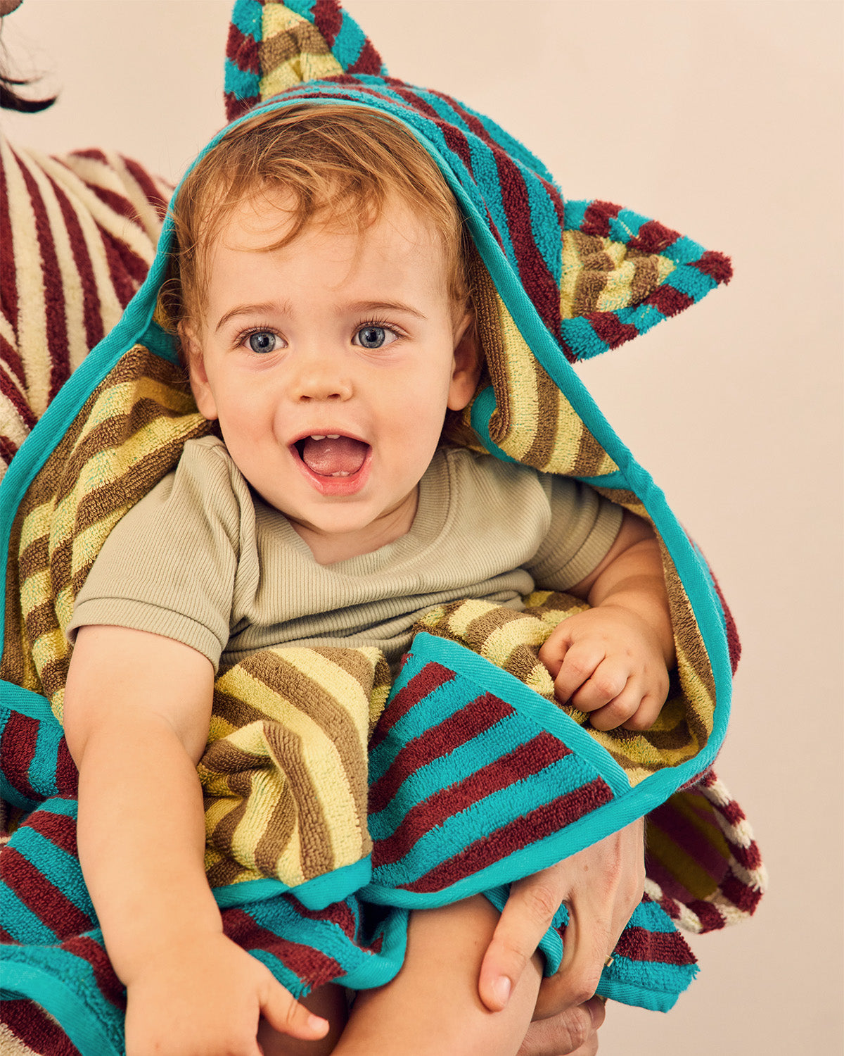 A baby wrapped in a colorful striped hooded towel with ears, held by an adult against a plain beige background. 